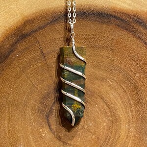 May include: A silver pendant necklace with a rectangular, multi-coloured stone wrapped in silver wire. The stone shows shades of green, brown, and red. The necklace is displayed on a wooden surface with a purple crystal background.