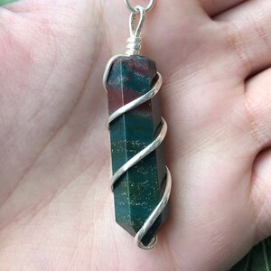 May include: A silver wire-wrapped pendant featuring a green, red, and brown jasper stone. The stone is a long, rectangular shape and is wrapped in silver wire in a spiral pattern.