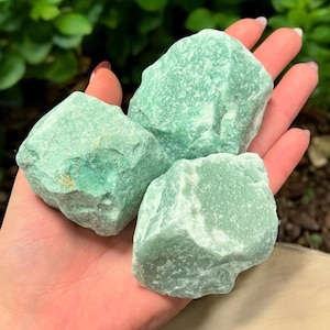 May include: Three rough-cut, light green aventurine stones. The stones are held in an open hand, with a blurred green background. The stones have a slightly textured surface and varying shades of green.