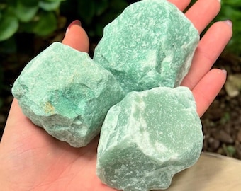 May include: Three rough-cut, light green aventurine stones. The stones are held in an open hand, with a blurred green background. The stones have a slightly textured surface and varying shades of green.