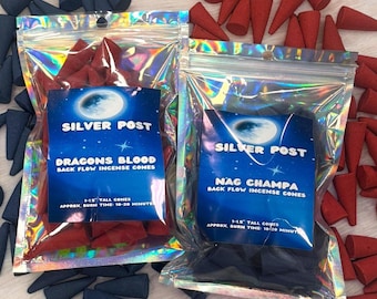 May include: Two bags of backflow incense cones. The bag on the left is labeled "Dragons Blood Back Flow Incense Cones" and the bag on the right is labeled "Nag Champa Back Flow Incense Cones". Both bags are silver with a holographic finish and feature a crescent moon and stars.