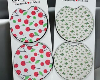 May include: Set of four car coasters. Two coasters feature a pattern of red cherries with green leaves on a white background. The other two have a pattern of green cacti on a white background. Each coaster has a curved cutout for easy removal. The text "Car Coasters" is printed on the top.