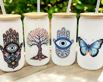 May include: Four white glass tumblers with bamboo lids and straws. Each tumbler features a different design: a Hamsa hand with an eye, a colorful tree, a Hamsa hand with an eye, and a blue butterfly. The tumblers are on a wooden surface.