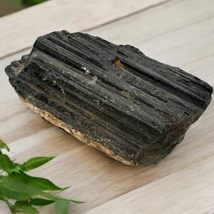 May include: A large, black, rough-hewn piece of black tourmaline, also known as schorl, with a striated surface. The stone is resting on a light wood surface.