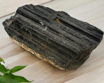 May include: A large, black, rough-hewn piece of black tourmaline, also known as schorl, with a striated surface. The stone is resting on a light wood surface.