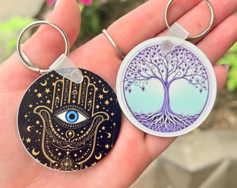 May include: Two round keychains. One features a black background with a gold Hamsa hand design and a blue eye. The other keychain has a purple tree design with roots and branches. Both keychains have silver key rings.