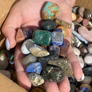 May include: A hand holding a variety of tumbled stones, including blue, green, brown, and red stones. The stones are smooth and polished.