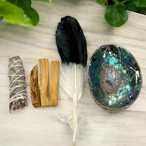 May include: An assortment of spiritual items including a white sage bundle, three palo santo sticks, a black and white feather, and an abalone shell bowl. The items are arranged on a light wood-grain surface.