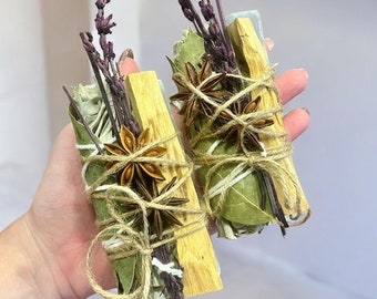 May include: Two bundles of dried herbs and wood for smudging, tied with twine. The bundles include sage, palo santo, lavender, and star anise.