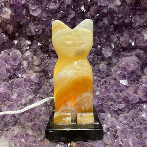 May include: A yellow onyx cat figurine lamp with a black base. The lamp is lit and the light shines through the onyx, creating a warm glow.