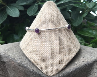 May include: A silver bangle bracelet with three amethyst gemstones. The bracelet is displayed on a beige fabric stand.