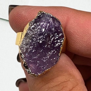 May include: A gold ring with a large, irregular shaped amethyst stone. The stone is a deep purple color and has a rough, natural texture.