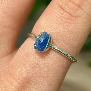 May include: A silver ring with a raw blue sapphire gemstone. The ring is on a person's finger.