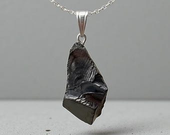 May include: A dark, irregularly shaped pendant made of a black, possibly obsidian-like material, suspended from a delicate silver chain. The pendant has a rough, textured surface and is displayed against a neutral gray background.