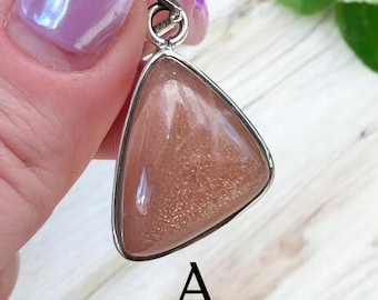 May include: A triangular pendant with a light brown sunstone gemstone. The pendant is set in silver and has a shiny, sparkly finish.
