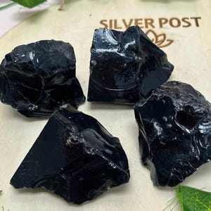 May include: Four pieces of raw, black obsidian stones. The stones have a glossy, reflective surface and irregular shapes. The image is set on a light-colored wooden surface with the text "SILVER POST" visible. Green leaves are in the background.