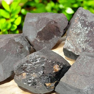 K&ouml;nnte beinhalten: Eine Sammlung dunkelgrauer H&auml;matitsteine mit metallischem Glanz. Die Steine haben raue, unregelm&auml;&szlig;ige Formen und unterschiedliche Gr&ouml;&szlig;en. Der Hintergrund zeigt verschwommenes gr&uuml;nes Laub und eine Holzoberfl&auml;che.