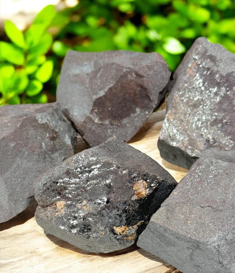 K&ouml;nnte beinhalten: Eine Sammlung dunkelgrauer H&auml;matitsteine mit metallischem Glanz. Die Steine haben raue, unregelm&auml;&szlig;ige Formen und unterschiedliche Gr&ouml;&szlig;en. Der Hintergrund zeigt verschwommenes gr&uuml;nes Laub und eine Holzoberfl&auml;che.