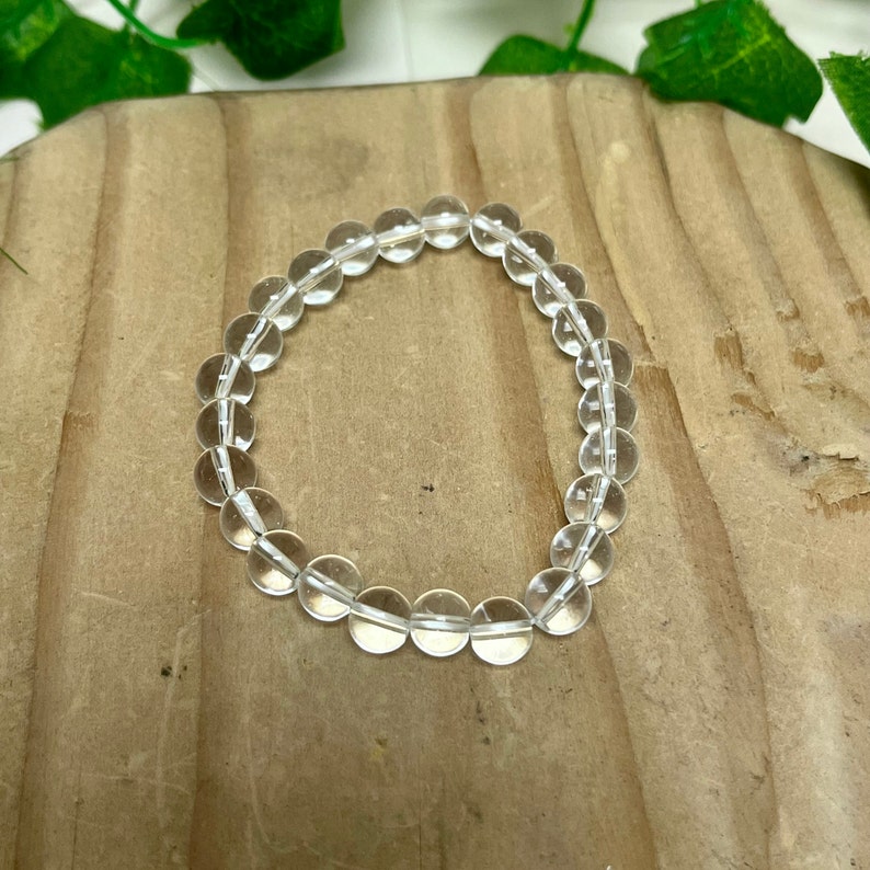 May include: A clear quartz crystal bead bracelet. The bracelet is made of round, transparent beads strung together. The beads are approximately 0.4 inches in diameter. The bracelet is displayed on a wooden surface.