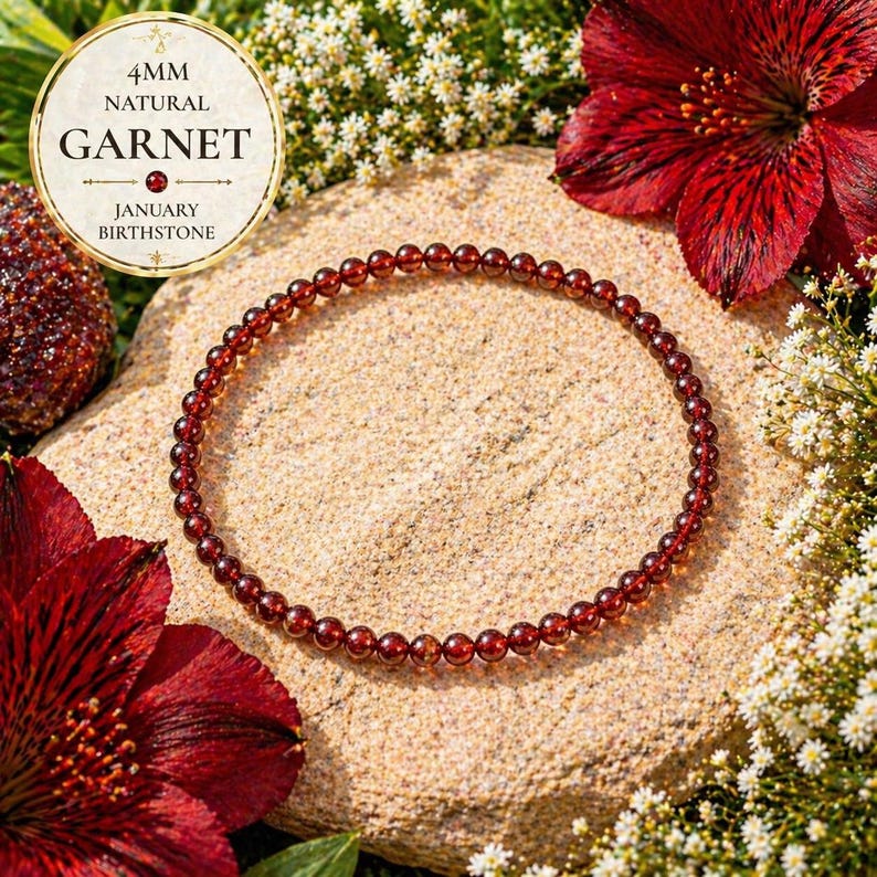 May include: A delicate bracelet made of small, round, deep red garnet beads. The bracelet is displayed on a textured, light brown surface, surrounded by red flowers and white blossoms. The text on the image reads "4mm Natural Garnet January Birthstone."