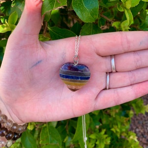 K&ouml;nnte beinhalten: Eine silberne Kettenkette mit einem herzf&ouml;rmigen Anh&auml;nger aus geschichteten Edelsteinen. Der Anh&auml;nger hat Streifen in den Farben Lila, Blau, Gelb und Braun.
