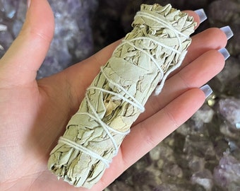 May include: A white sage smudge stick wrapped in twine, ready for burning. The stick is held in a hand, with a background of purple and white crystals.