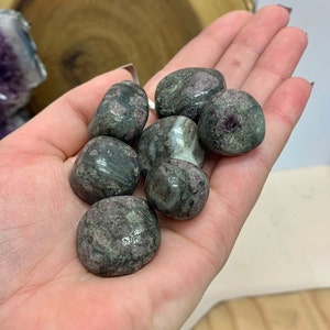 May include: A collection of tumbled green and purple stones with a speckled pattern. The stones are smooth and polished.