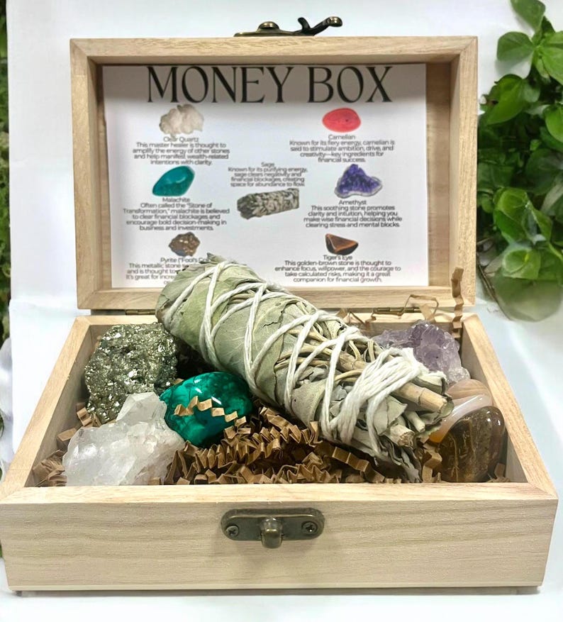 Peut inclure: Bo&icirc;te en bois avec un couvercle sur lequel est inscrit "MONEY BOX". La bo&icirc;te est ouverte et contient une vari&eacute;t&eacute; de cristaux, un b&acirc;tonnet de sauge et un morceau de papier avec des informations sur les cristaux.