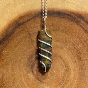May include: A green and brown gemstone pendant with a silver wire wrap, suspended from a silver chain. The pendant is rectangular with a pointed bottom. The background is a wooden surface.