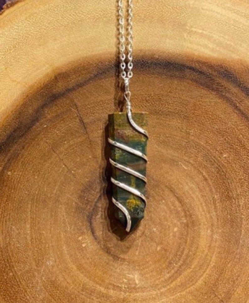 May include: A green and brown gemstone pendant with a silver wire wrap, suspended from a silver chain. The pendant is rectangular with a pointed bottom. The background is a wooden surface.
