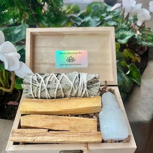 May include: A wooden box with a lid open, revealing a home cleansing kit. The kit includes sage, palo santo, and selenite. The box has a label that reads "Home Cleansing Kit" with a symbol of a flower.