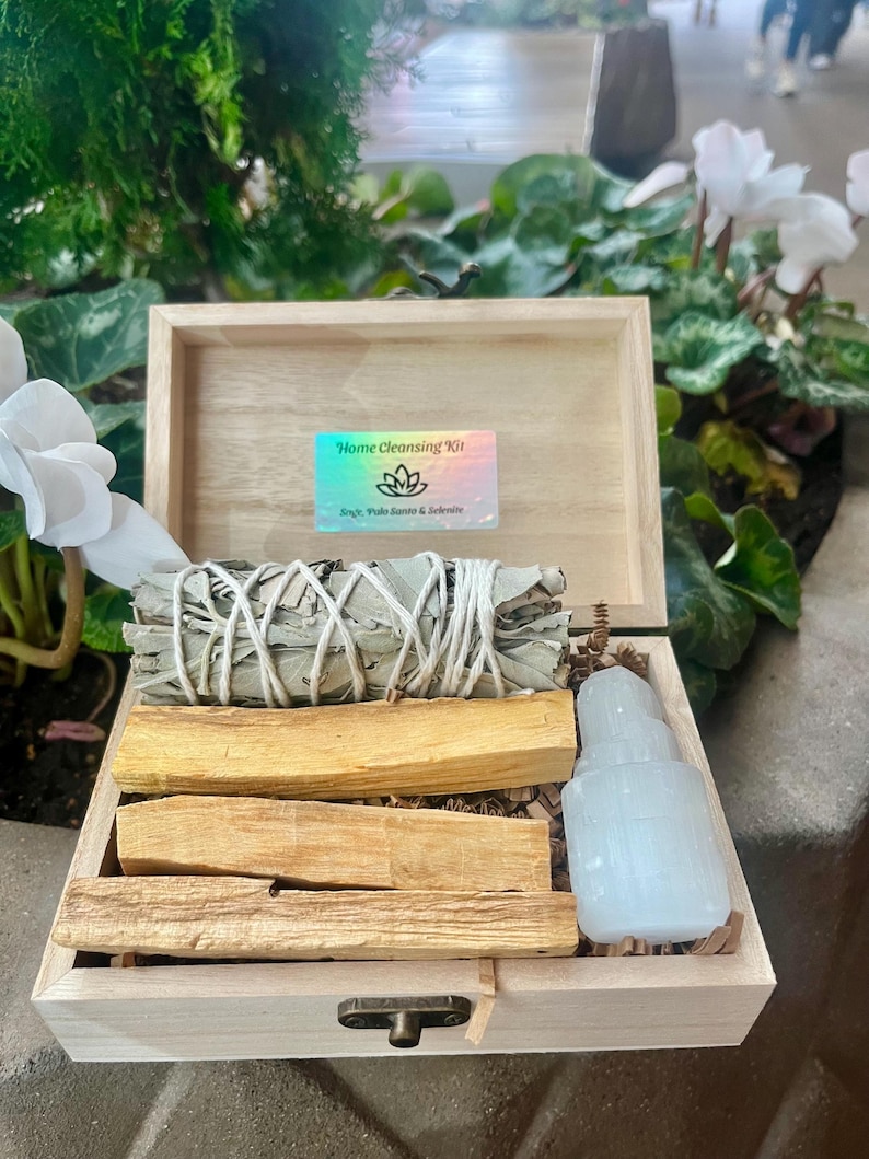May include: A wooden box with a lid open, revealing a home cleansing kit. The kit includes sage, palo santo, and selenite. The box has a label that reads "Home Cleansing Kit" with a symbol of a flower.