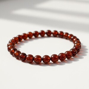 May include: A beaded bracelet featuring round, reddish-brown beads. The beads have a speckled appearance and are strung together to form a circular shape. The bracelet is displayed on a white surface, with soft shadows.