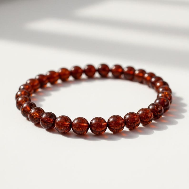 May include: A beaded bracelet featuring round, reddish-brown beads. The beads have a speckled appearance and are strung together to form a circular shape. The bracelet is displayed on a white surface, with soft shadows.