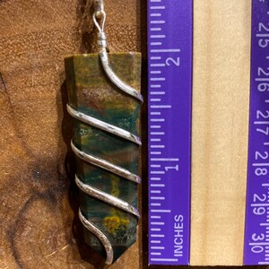 May include: A gemstone pendant necklace. The pendant is a rectangular, multi-coloured stone wrapped in silver wire, approximately 5 cm long. The stone features green, brown, and red hues. The necklace chain is silver.
