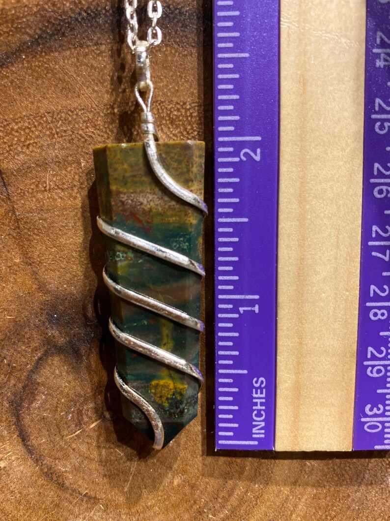 May include: A gemstone pendant necklace. The pendant is a rectangular, multi-coloured stone wrapped in silver wire, approximately 5 cm long. The stone features green, brown, and red hues. The necklace chain is silver.