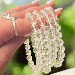 May include: Clear quartz crystal bead bracelets. The bracelets are made of transparent, round beads. The beads are strung together on an elastic band. The bracelets are held up against a blurred green background.