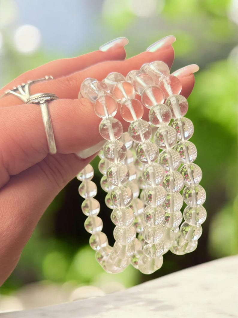 May include: Clear quartz crystal bead bracelets. The bracelets are made of transparent, round beads. The beads are strung together on an elastic band. The bracelets are held up against a blurred green background.