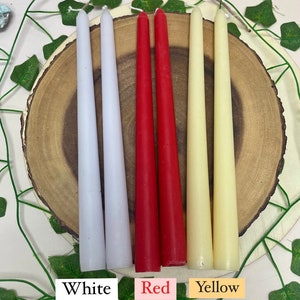 May include: Three pairs of taper candles in white, red, and yellow. The candles are arranged on a wooden surface.