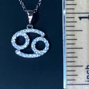 May include: Silver pendant necklace featuring a Cancer zodiac sign with clear cubic zirconia stones.