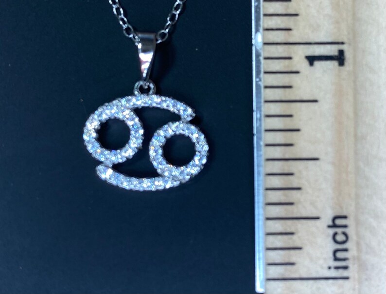 May include: Silver pendant necklace featuring a Cancer zodiac sign with clear cubic zirconia stones.