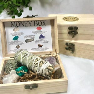 Peut inclure: Bo&icirc;te en bois avec un couvercle &agrave; charni&egrave;res, &eacute;tiquet&eacute;e "Money Box". La bo&icirc;te contient divers cristaux et un fagot de sauge, notamment du quartz clair, de la cornaline, de la malachite, de l'am&eacute;thyste, de l'&oelig;il de tigre et de la pyrite. La bo&icirc;te comprend &eacute;galement une carte avec des descriptions de chaque cristal et de ses propri&eacute;t&eacute;s.