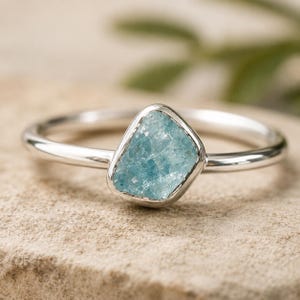 May include: A silver ring featuring a single, rough-cut, light blue gemstone set in a smooth metal bezel. The band is polished and circular, holding the irregular, textured stone securely in the center of the setting for a clean, simple look.