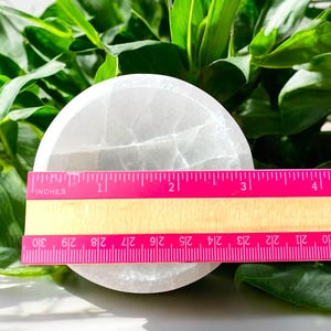 May include: A circular, white selenite crystal plate, measuring approximately 7.6 cm in diameter, with a pink ruler showing the measurement. The plate is set against a backdrop of green foliage.