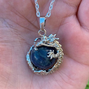 May include: Silver pendant necklace with a dark blue orb encased in a detailed dragon design. The dragon's head and claws are visible, adding intricate detail. The necklace has a delicate chain.