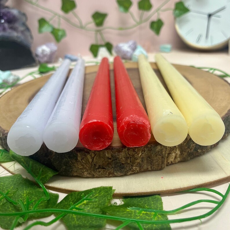May include: Five taper candles in various colors, including white, red, and cream, arranged on a wooden surface. The candles are all the same size and shape, and they are all lit.