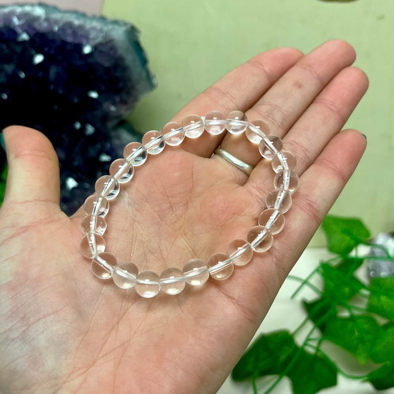 May include: A clear quartz crystal beaded bracelet. The bracelet is made of round, translucent beads strung together on a clear elastic cord. The bracelet is shown on a person's hand.