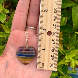 K&ouml;nnte beinhalten: Eine silberne Kettenkette mit einem herzf&ouml;rmigen Anh&auml;nger aus geschichteten Steinen in den Farben Lila, Blau, Gelb und Braun. Der Anh&auml;nger hat einen Durchmesser von etwa 2,5 cm.