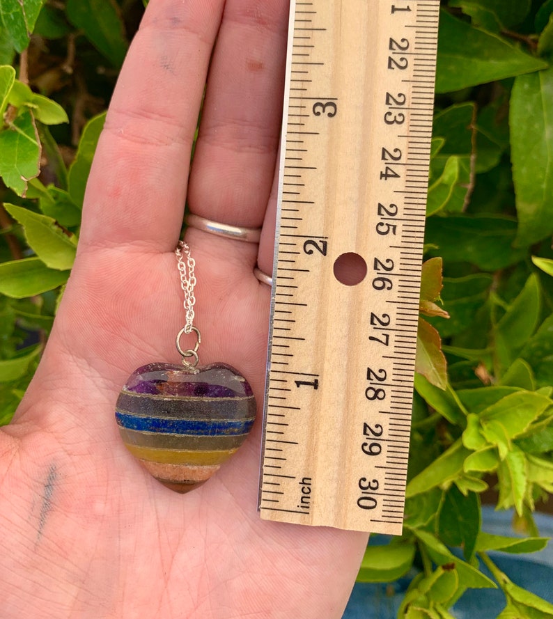 K&ouml;nnte beinhalten: Eine silberne Kettenkette mit einem herzf&ouml;rmigen Anh&auml;nger aus geschichteten Steinen in den Farben Lila, Blau, Gelb und Braun. Der Anh&auml;nger hat einen Durchmesser von etwa 2,5 cm.