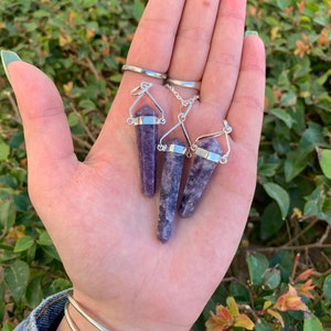 May include: Three purple amethyst crystal point pendants on silver chains. The pendants are hanging from a hand.